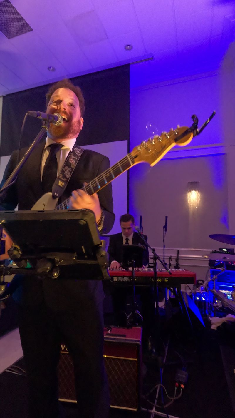 Dave Menzo singing and playing guitar, the keys player behind him