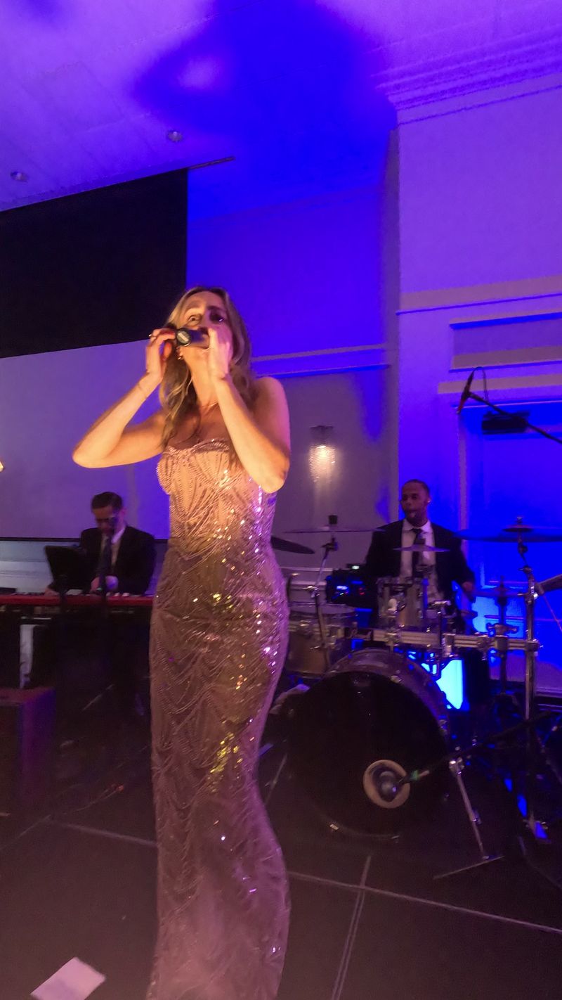 The singer at the microphone in a gold sequined gown, keys and drums lit purple behind her